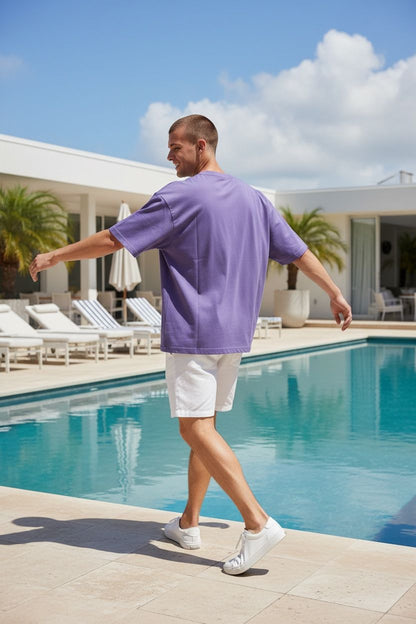 Man in a purple shirt and white shorts walking by a pool with palm trees and lounge chairs in the background.